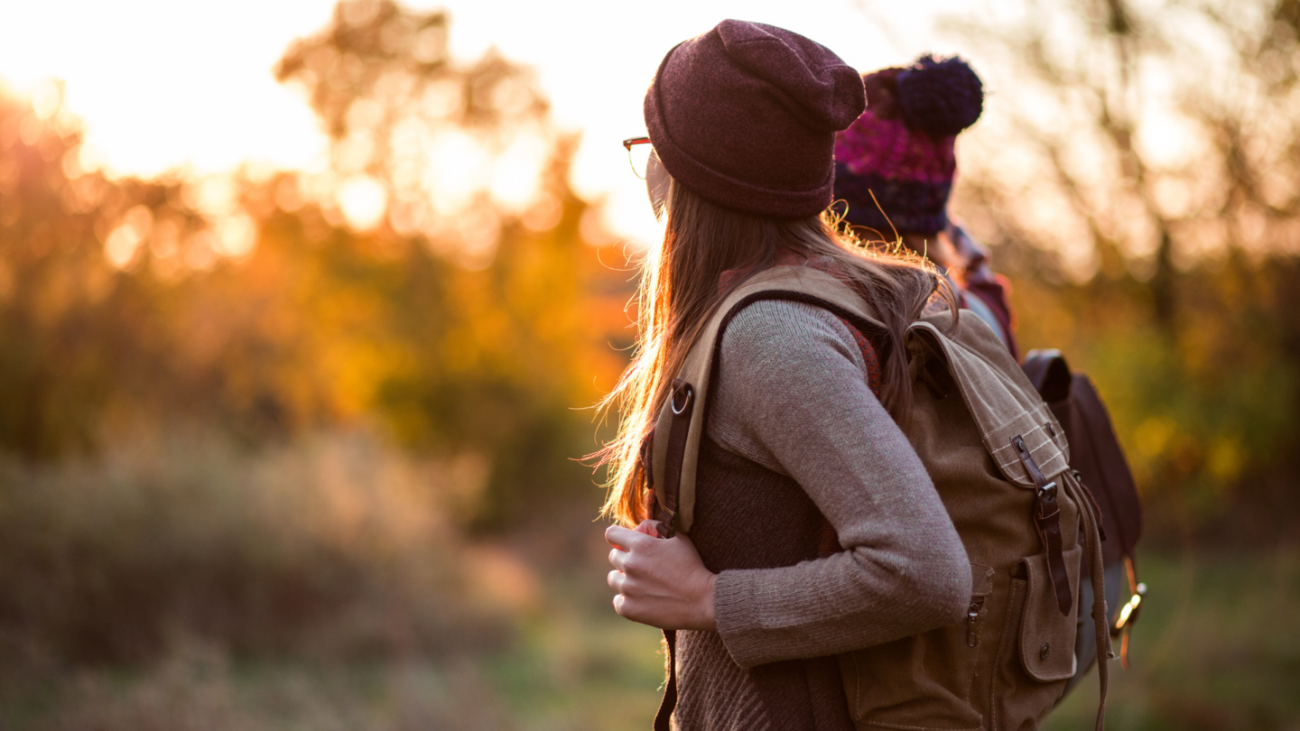 Zwei Wanderer in warmer Herbstkleidung spazieren durch die farbenfrohe Natur rund um den Tegernsee. Die goldenen Laubfarben und das sanfte Licht des Sonnenuntergangs schaffen eine stimmungsvolle Kulisse